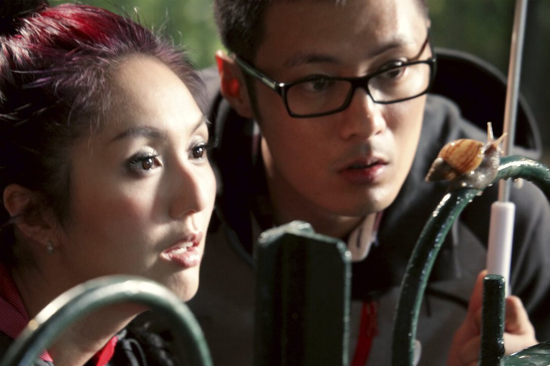 Two people observe a snail crawling on a railing, outdoors with blurred greenery in the background.