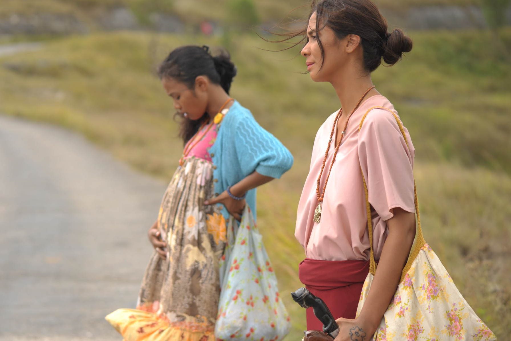 Two women stand on a rural path, each holding colorful bags with determined expressions. The grassy landscape stretches into the distance.