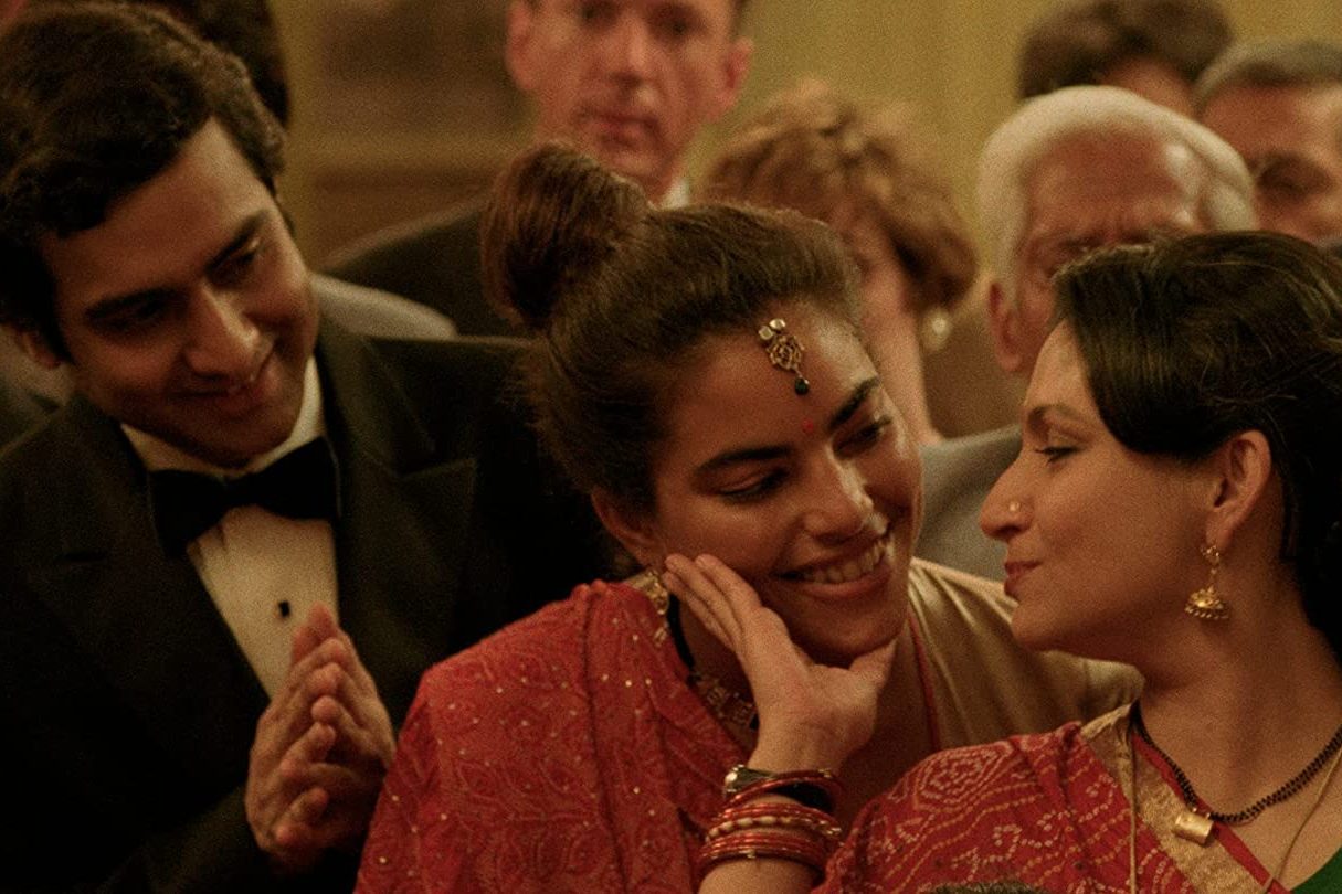 Two women in traditional attire share a tender moment while a man in a tuxedo smiles in the background at a social gathering.