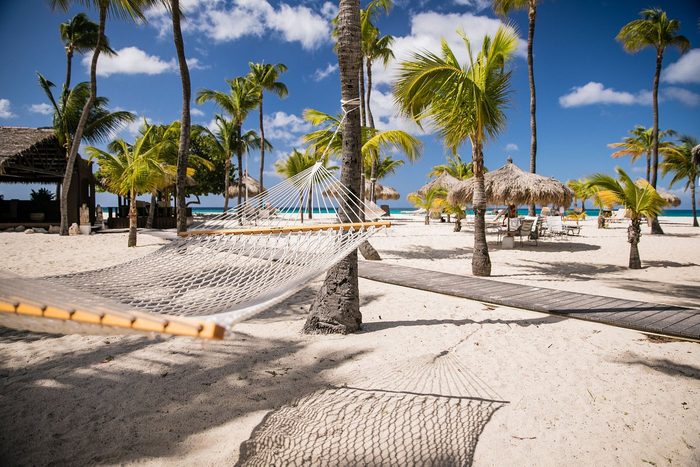 Hammock hangs between palm trees on a sunny beach, with thatched umbrellas and lounge chairs in the background under a clear blue sky.