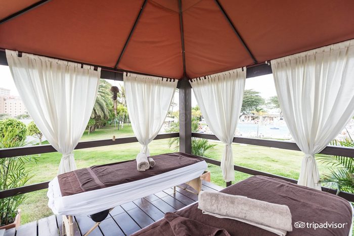 Massage tables lie under a gazebo with white curtains overlooking a grassy area beside a pool. Nearby are palm trees and a few people.