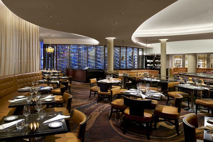 Elegant restaurant set for dining, featuring plush brown seating and a striking wall of illuminated wine bottles, exuding a sophisticated ambiance.
