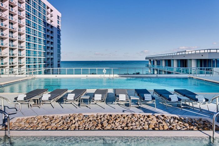 Loungers align poolside under a clear sky, flanked by modern buildings, overlooking a serene ocean view.