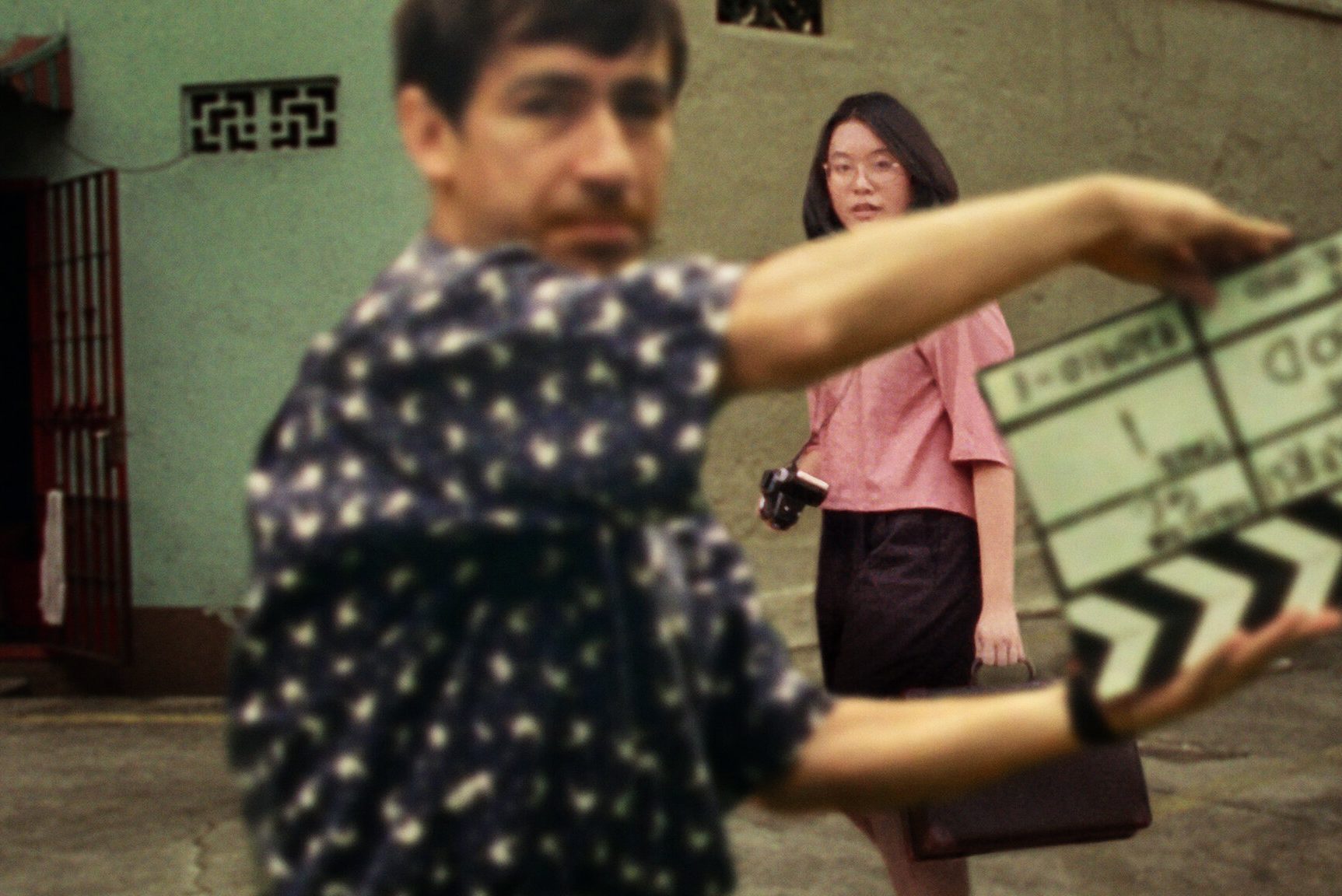 Clapperboard in motion, held by a person, with a woman carrying a camera and briefcase in the background on a street.