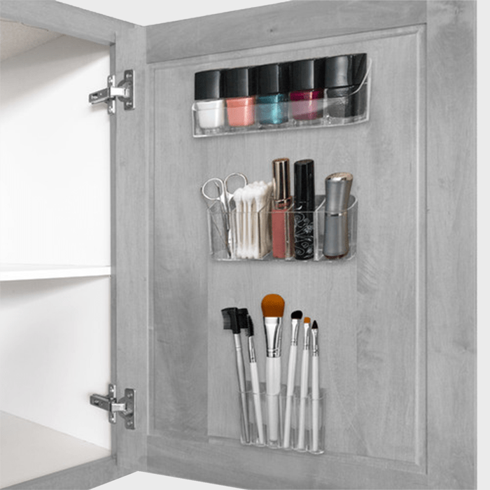 Makeup supplies are organized in clear holders on a cabinet door. Context is an open bathroom cabinet with shelves.