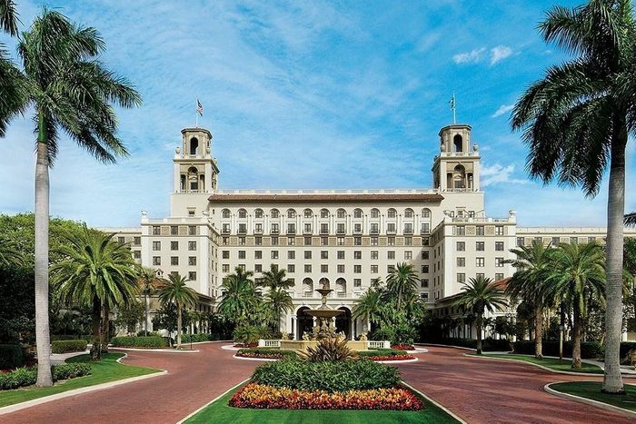 Grand building stands amid lush palm trees, with red-brick pathways leading to a central fountain under a bright blue sky.