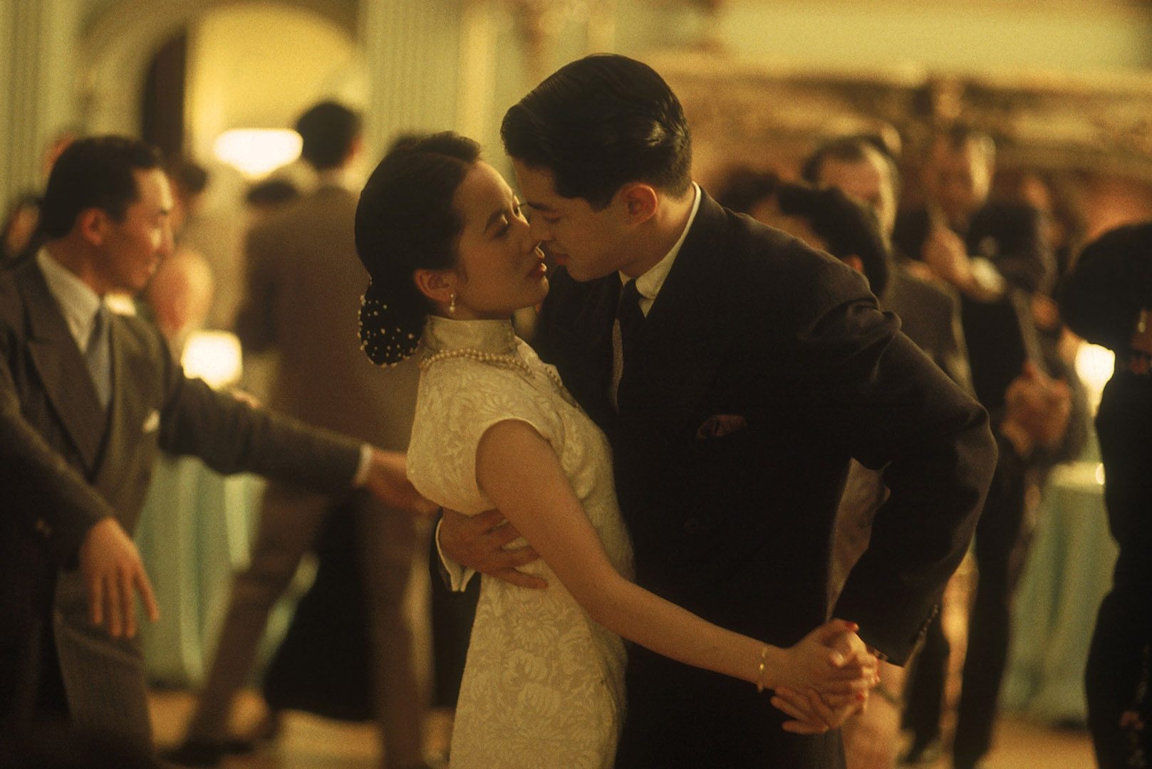 A couple dances closely, elegantly dressed, in a dimly lit, crowded ballroom with people in the background.