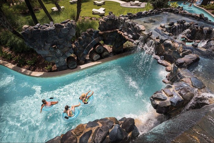 People float in a lazy river beneath rock structures, surrounded by lush greenery and lounge areas.