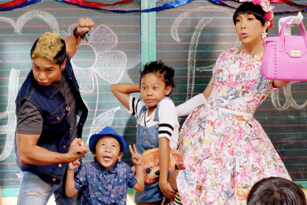 People pose energetically; one flexes muscles, another holds a plush toy, while someone in a floral dress displays a pink handbag. They're against a chalkboard.