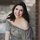 A person smiles, sitting on a brick ledge, surrounded by dappled sunlight on a paved ground.