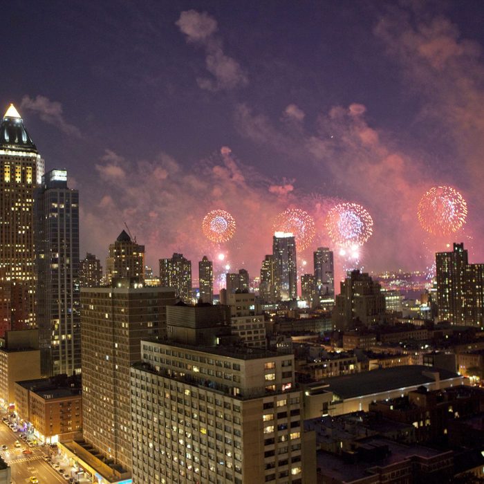 Fireworks explode over a city skyline at dusk, with tall buildings illuminated below and a vibrant, smoky sky above.