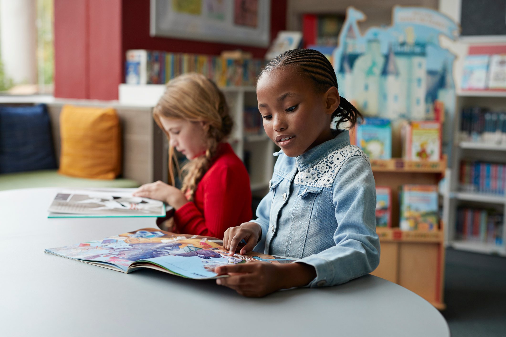 Two children read picture books at a table in a colorful library setting.