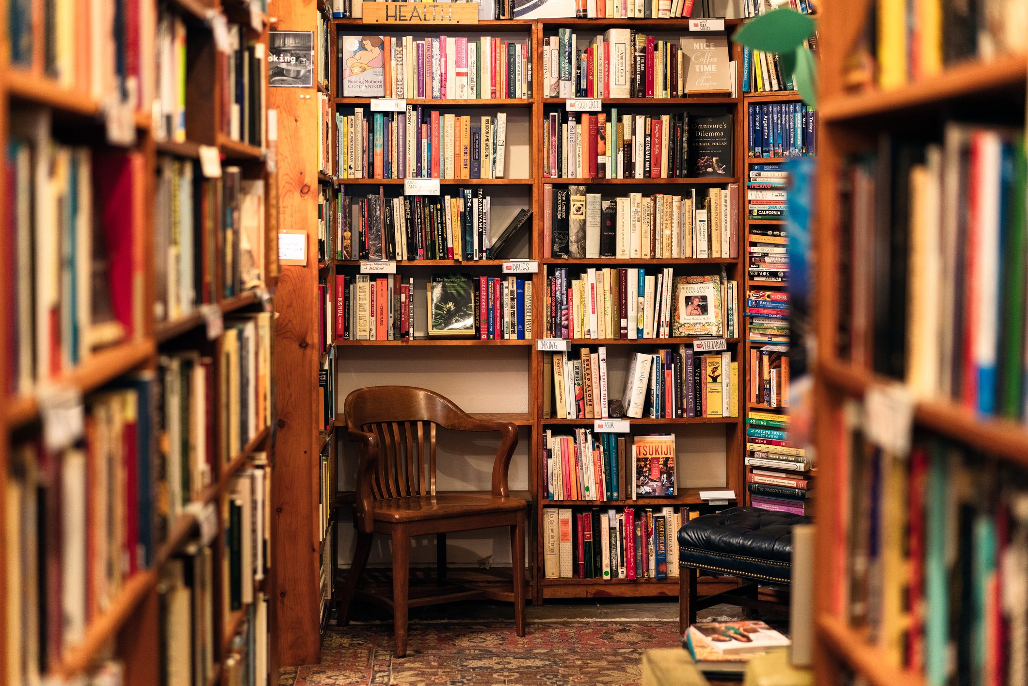 Bookshelves display colorful books, surrounding a wooden chair in a cozy, carpeted library with warm lighting and wooden furniture.
