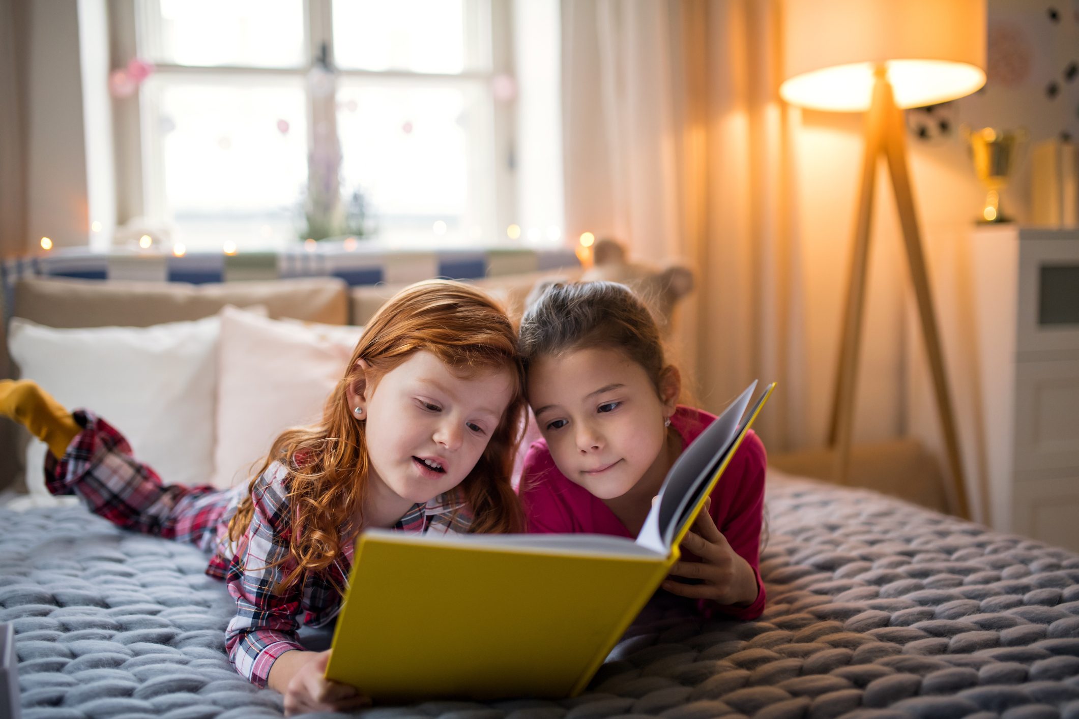 Two children read a yellow book on a bed in a cozy, softly-lit bedroom with pillows and a floor lamp.