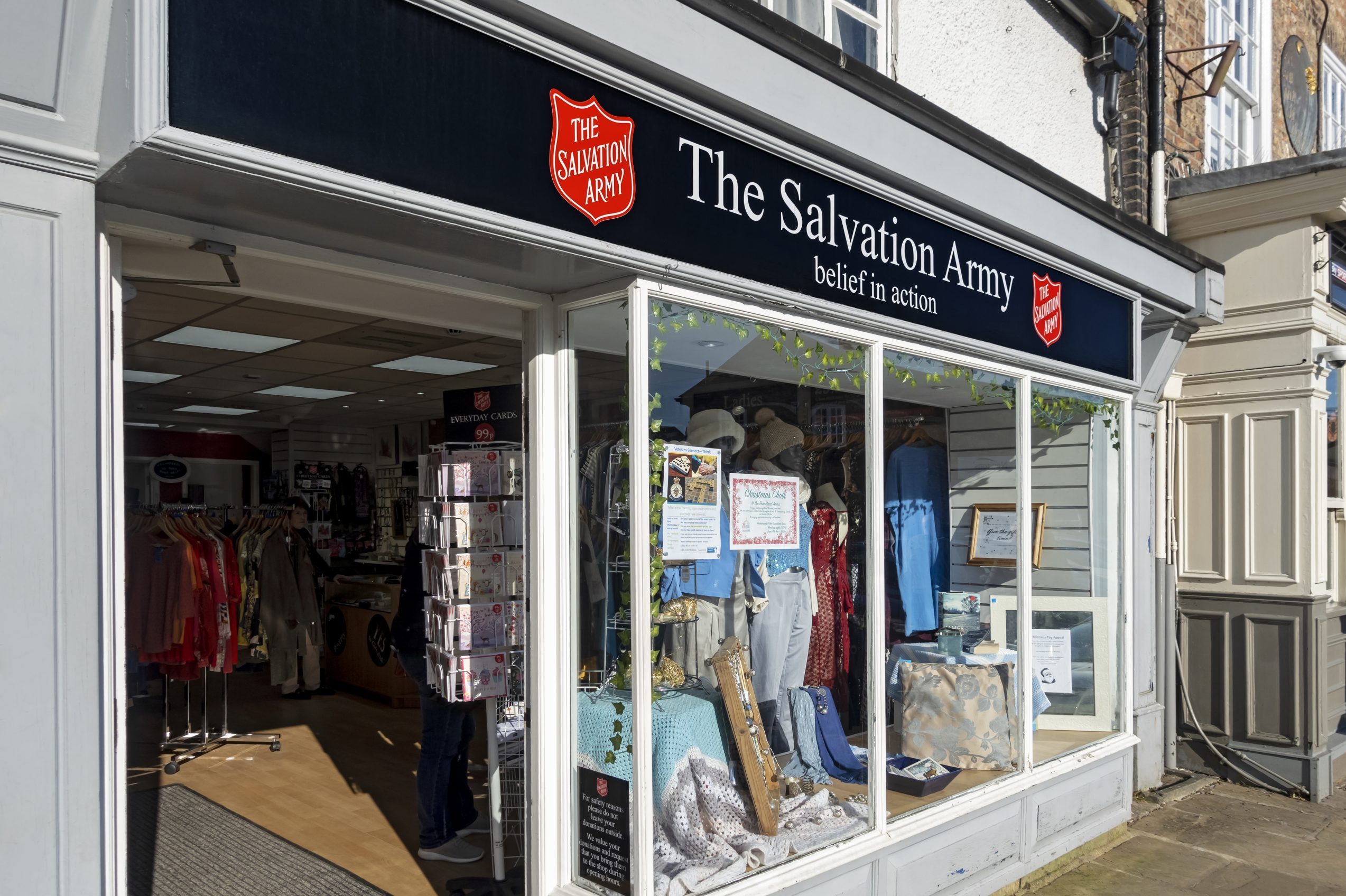 Storefront displays clothing and cards; interior visible through windows. Sign reads: "The Salvation Army â€“ belief in action." Sunlight illuminates entrance.