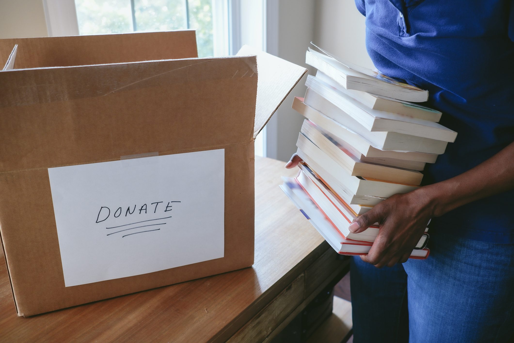 A person carries books toward an open cardboard box labeled "DONATE" on a wooden table near a window.