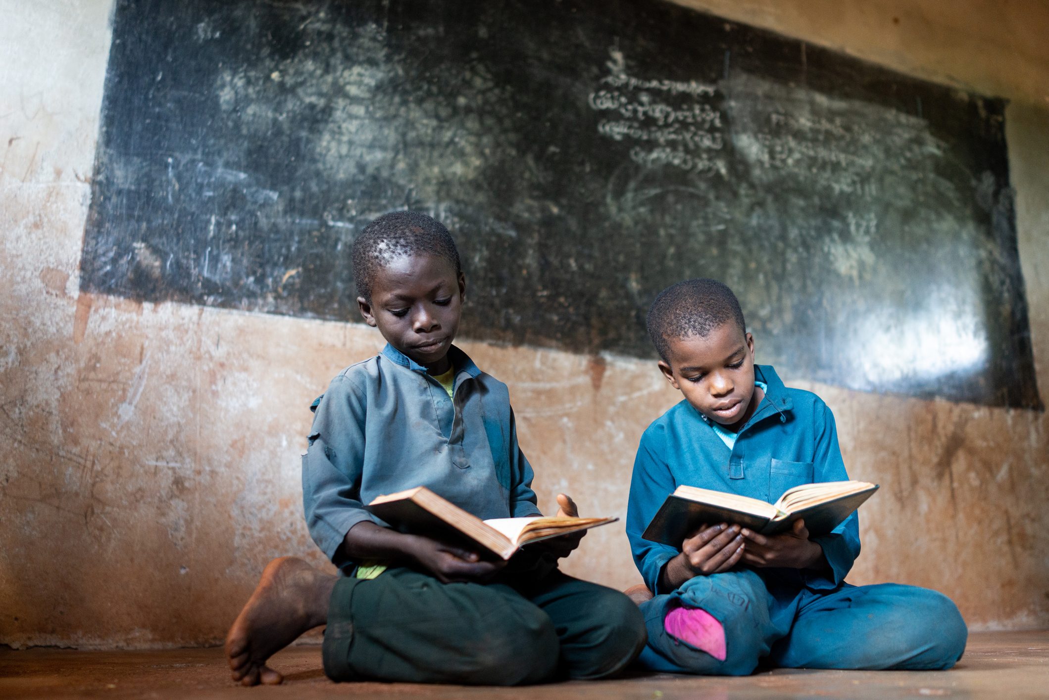 Two boys read books while sitting on the floor, in front of a chalkboard, in a dimly lit classroom.