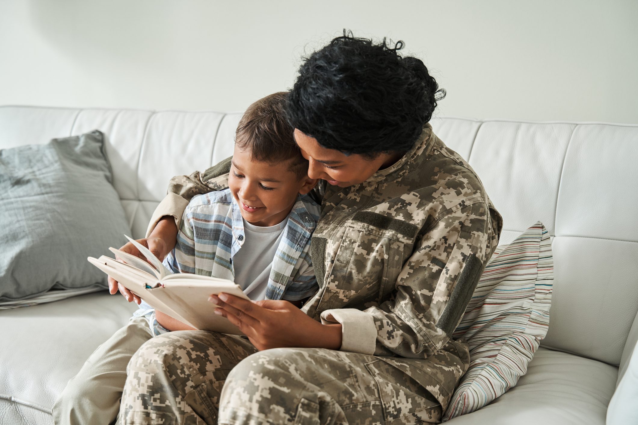 Child and adult read a book together on a white sofa, with the adult wearing a military uniform and colorful pillows in the background.
