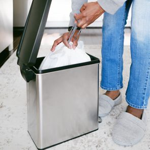 Hands pull a white trash bag from a stainless steel bin, surrounded by a kitchen setting with white cabinets and a light-patterned rug.