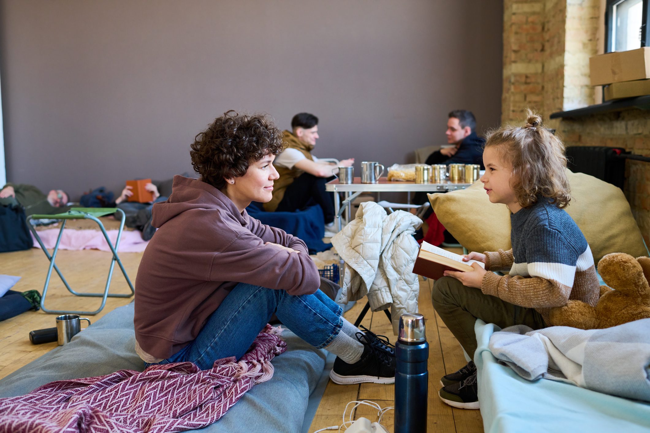 Two people sit and converse in a cozy room with blankets and cushions, as others relax or read in the background.