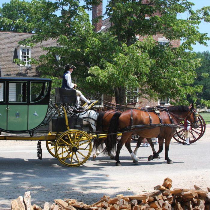 Horse-drawn carriage moves along a street, driven by a person in historical attire, with trees and a brick building in the background.