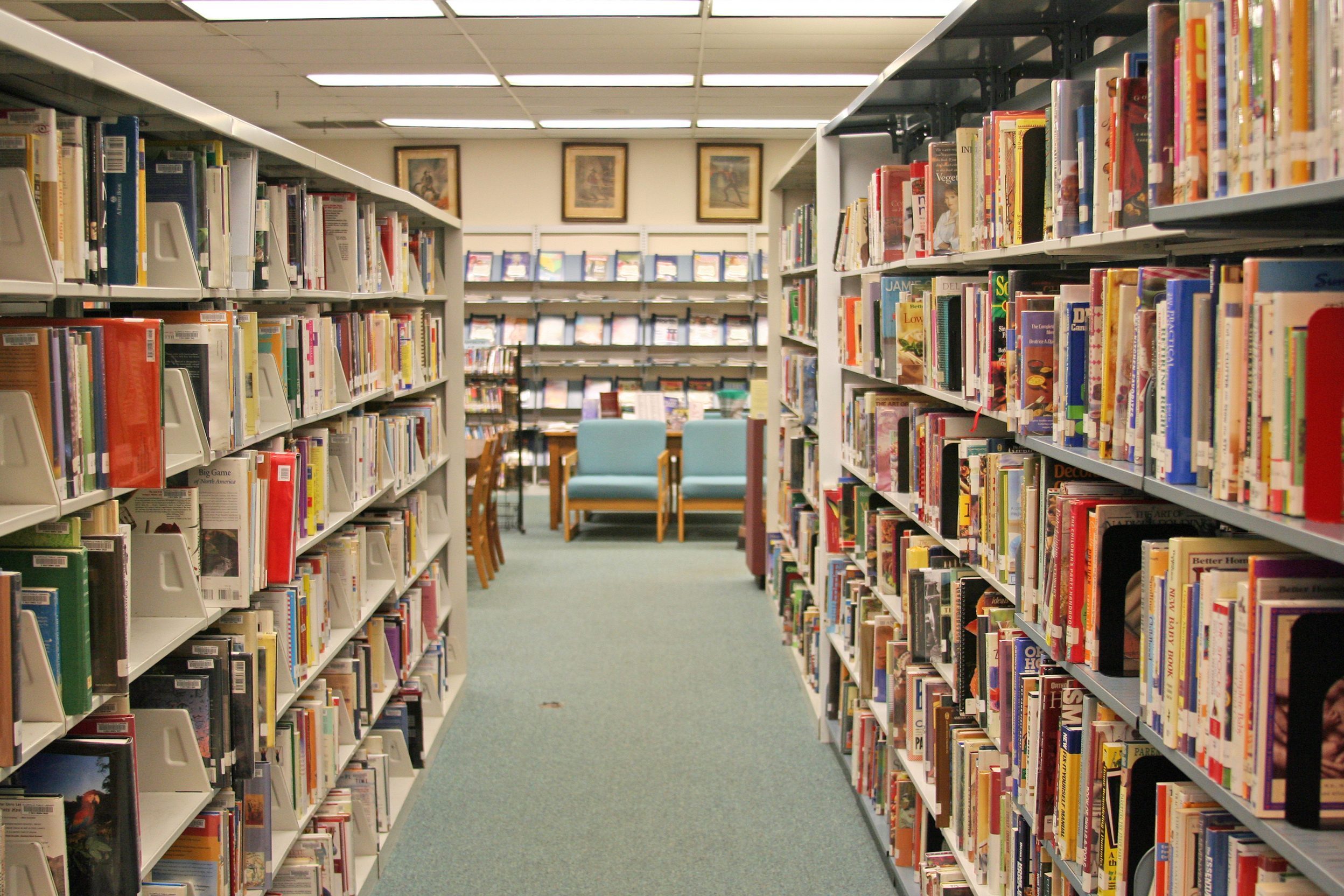 Books sit on shelves lining a library aisle, leading to a reading area with chairs and a table, under fluorescent lighting.