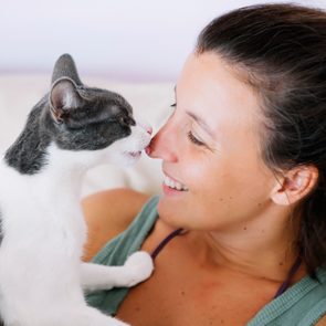 A cat gently touches a woman's nose with its paw while she smiles, both relaxing on a light-colored couch.