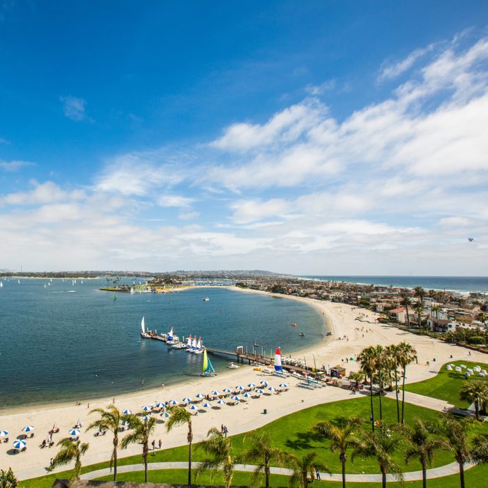 Beach curves around a bay with sailboats, surrounded by grassy park, palm trees, and beach umbrellas under a blue sky with scattered clouds.