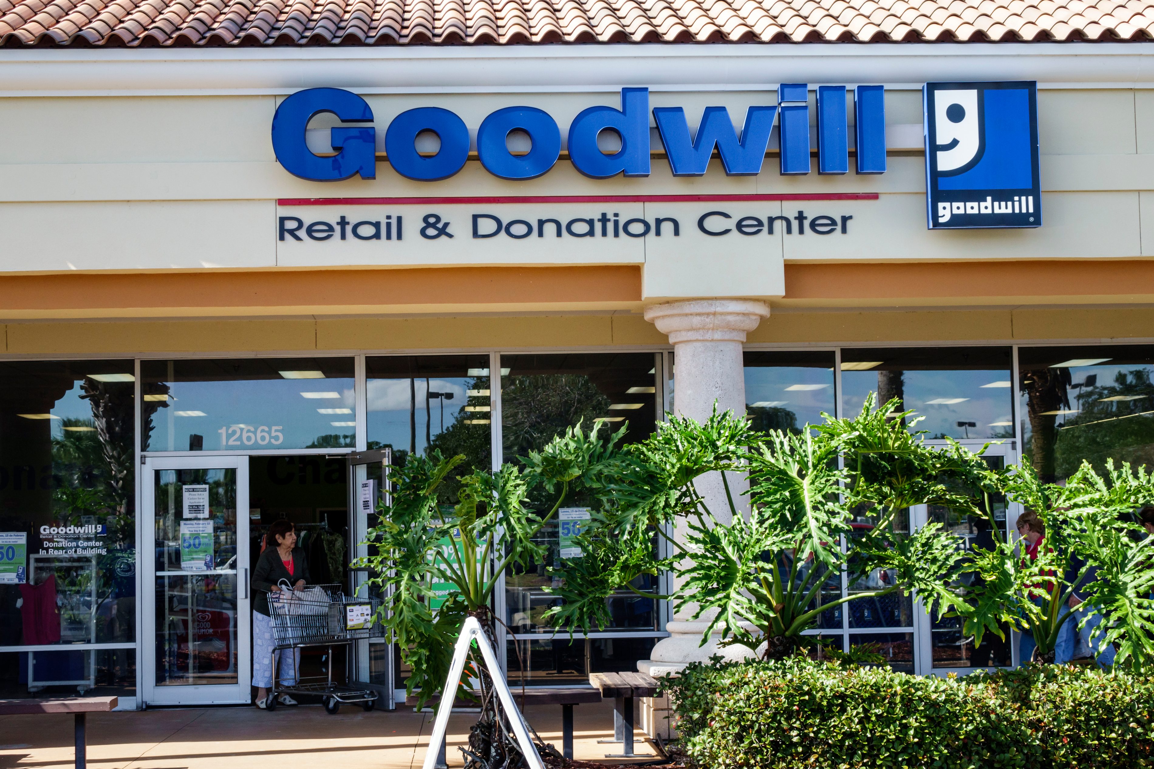 Goodwill store entrance; a person pushes a shopping cart out. Surrounding foliage and store signage are visible above.