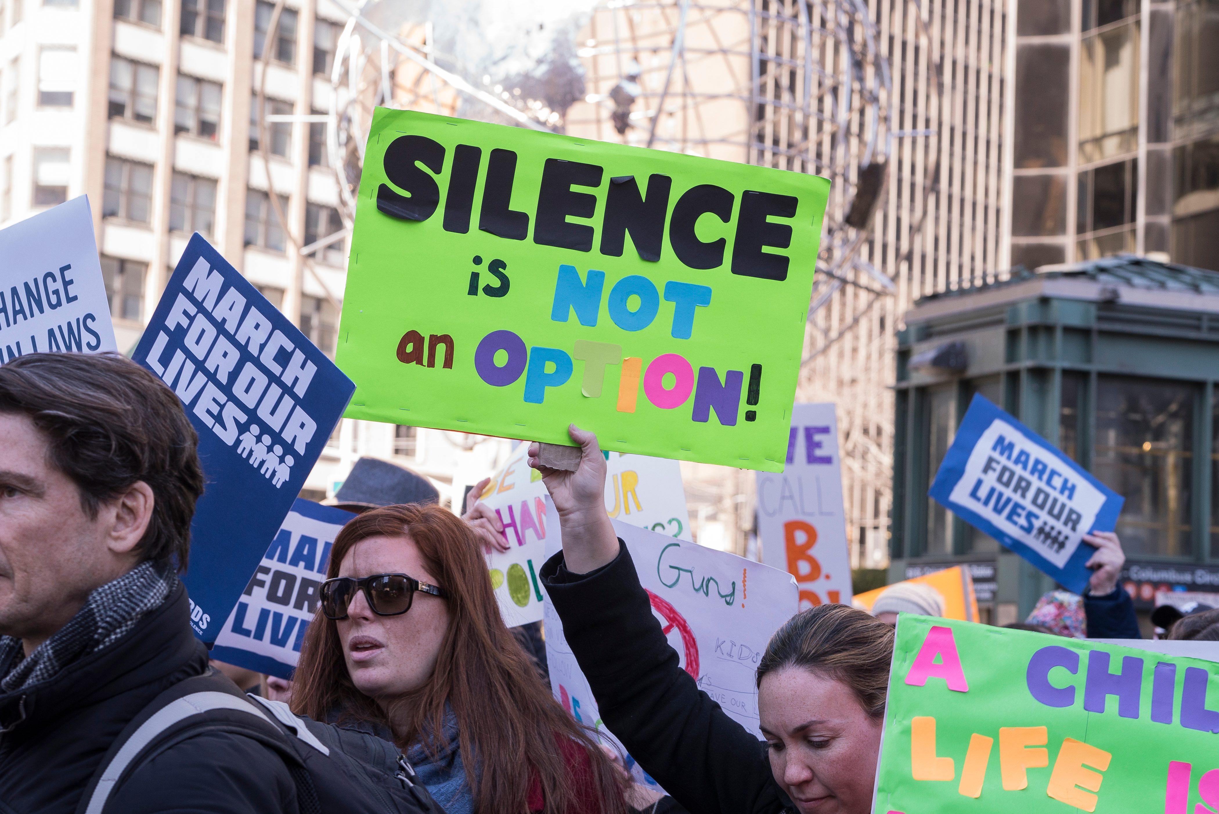 Protesters hold signs saying "Silence is Not an Option" and "March For Our Lives" in an urban setting.