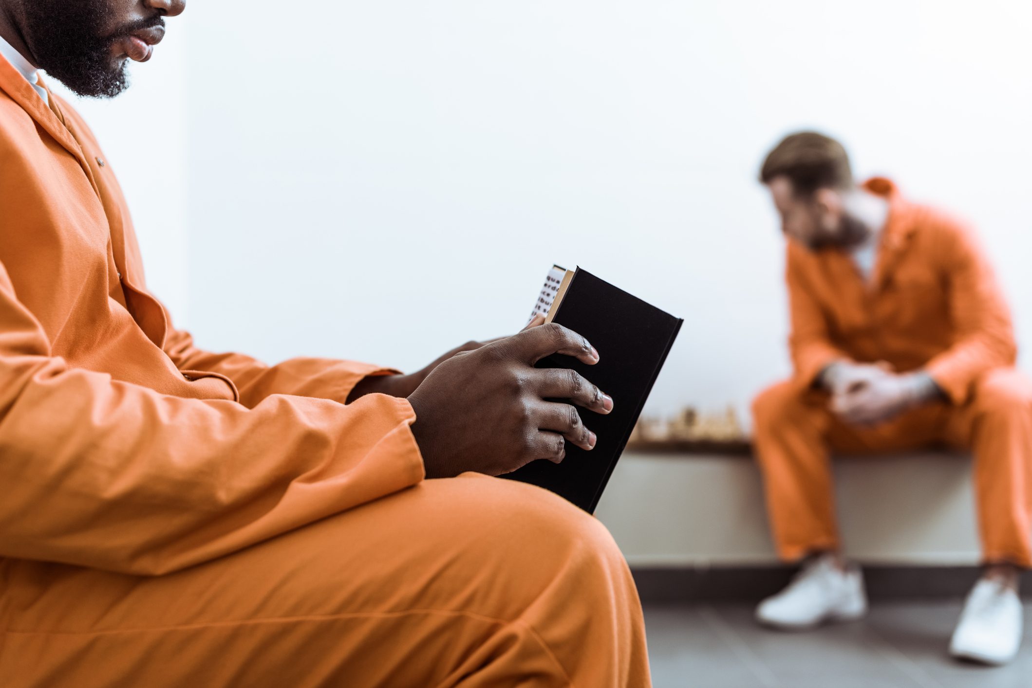 Person holds book, reading; another sits in background, both wearing orange jumpsuits, in a sparse room.