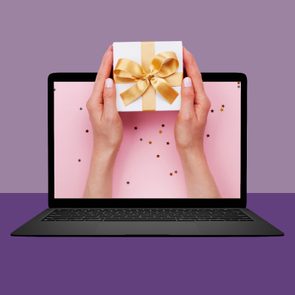 Hands hold a gift box with a gold ribbon, emerging from a laptop screen on a pink background with scattered star confetti.