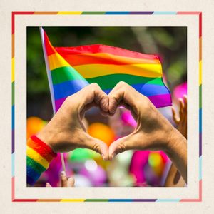 Hands form heart, framing a rainbow flag waving at a colorful outdoor event.
