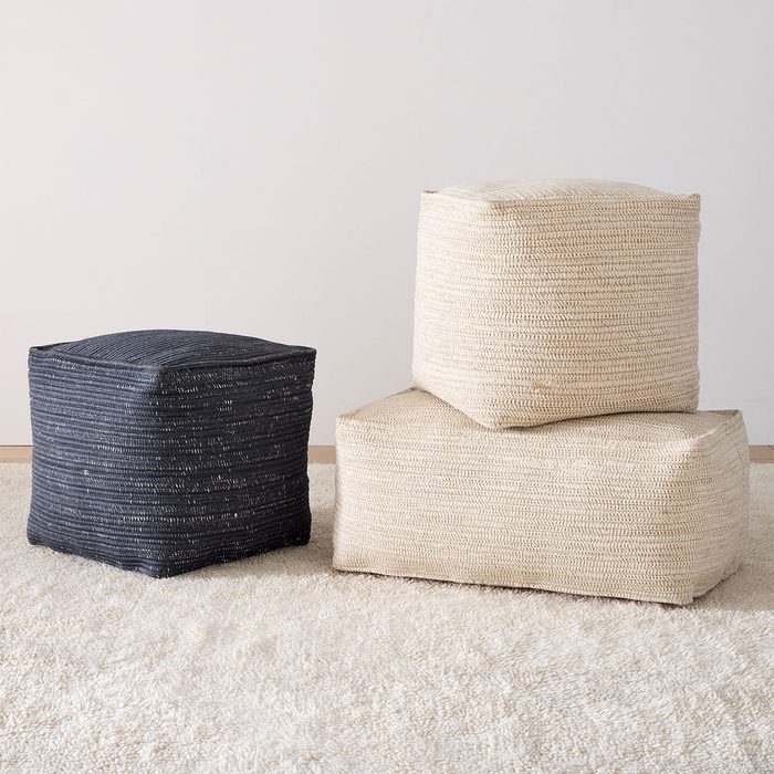 Three woven poufs are stacked and arranged on a light, textured carpet in a minimalist room setting.
