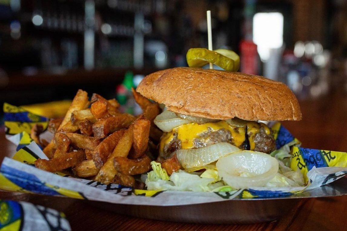 Burger with cheese and onions sits on a tray with fries, garnished by pickles and surrounded by a casual dining setting.