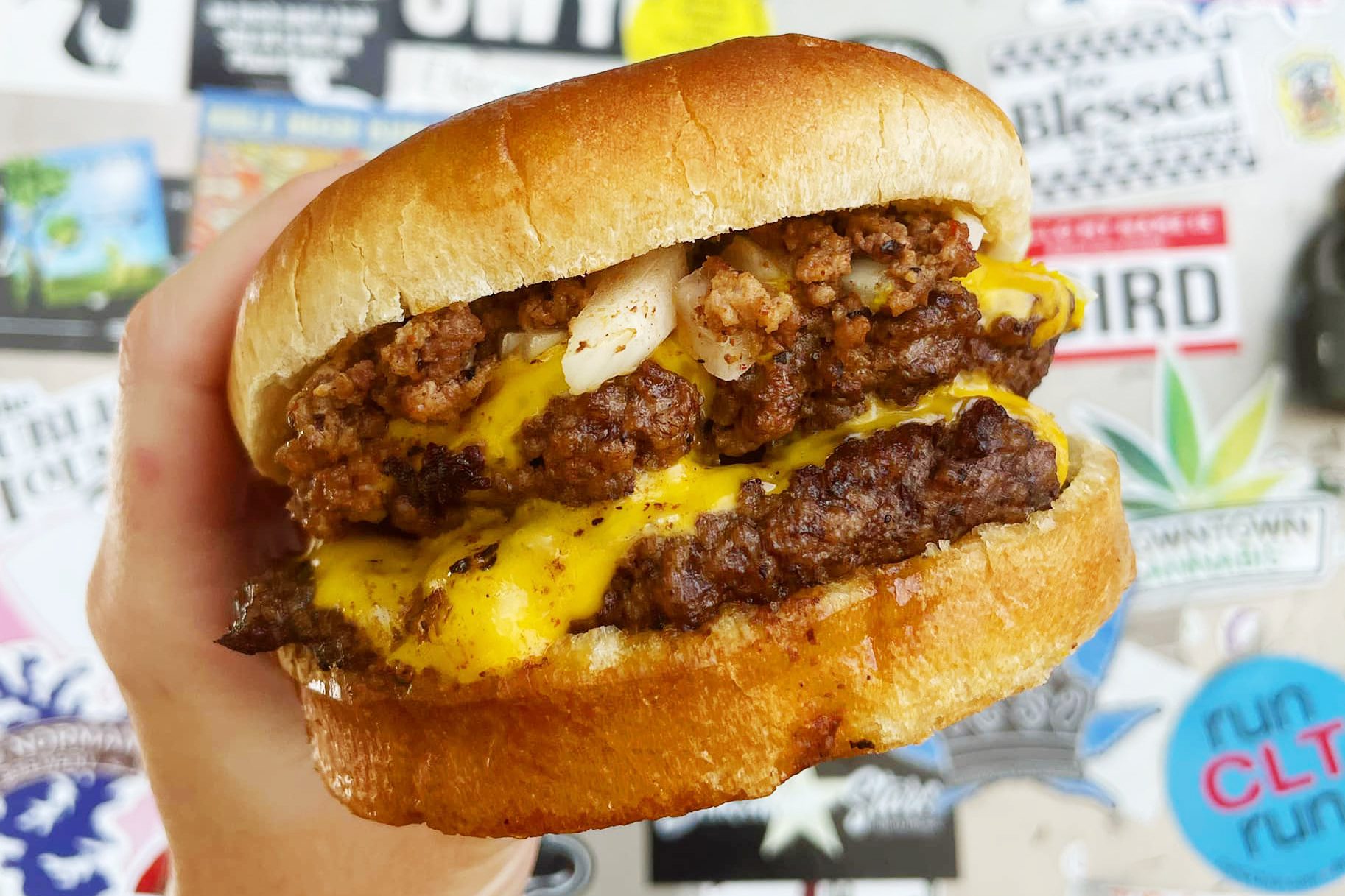 Burger held with a hand, showcasing melty cheese and crumbled meat. Background features various colorful stickers and signs.