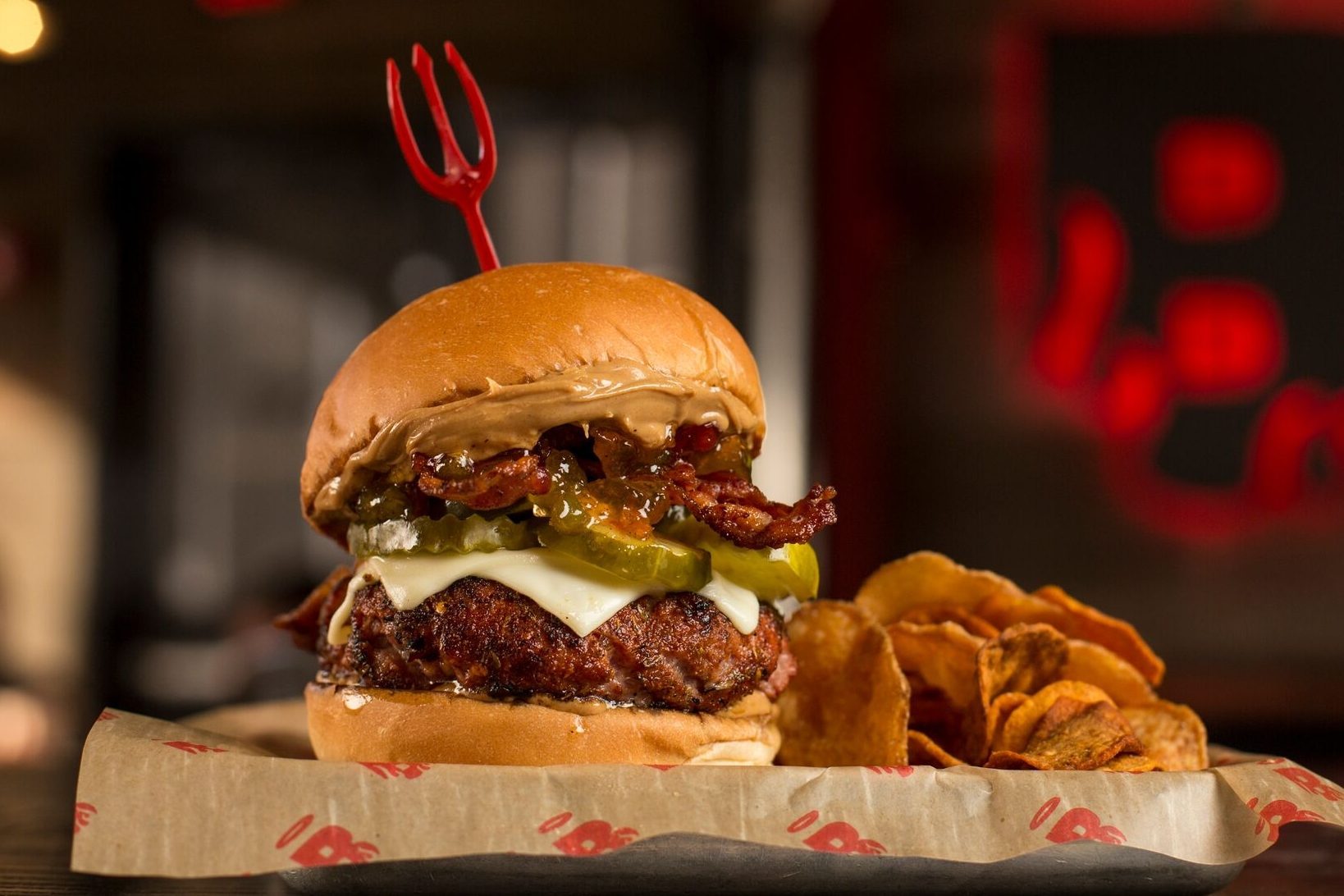 Burger stacked with pickles, bacon, and cheese, secured by a red pick, sits on paper. Chips are beside it in a warmly lit setting.