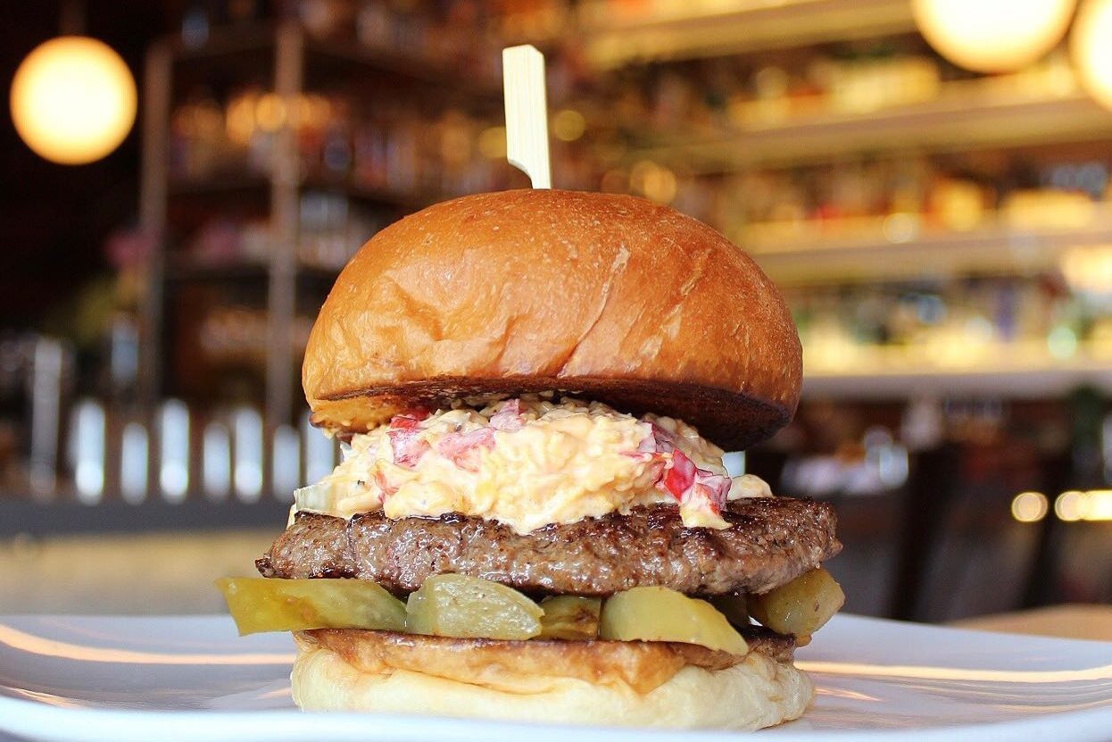 Burger stacked with pickles, cheese, and a beef patty sits on a plate in a restaurant with shelves in the background.