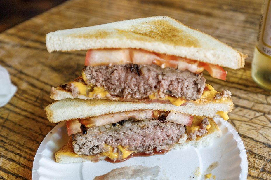 Toasted sandwich with beef patty, tomatoes, and cheese sits sliced on a white paper plate, placed on a wooden table.