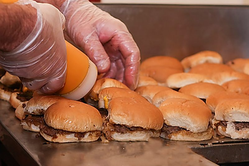 Gloved hands squeeze mustard onto small burgers on a grill.