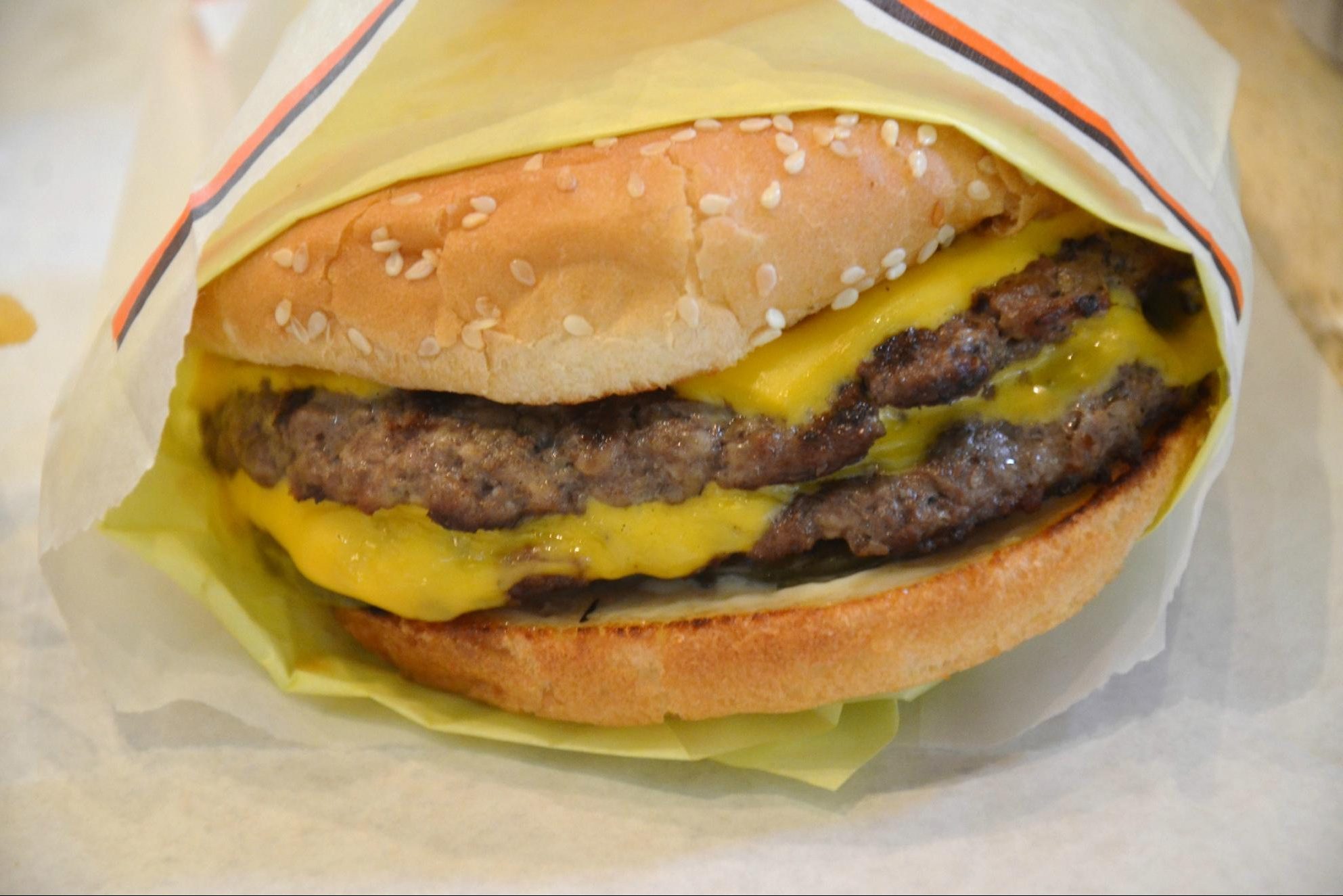 A double cheeseburger partially wrapped in paper sits on a surface, with sesame seed bun and melted cheese visible.