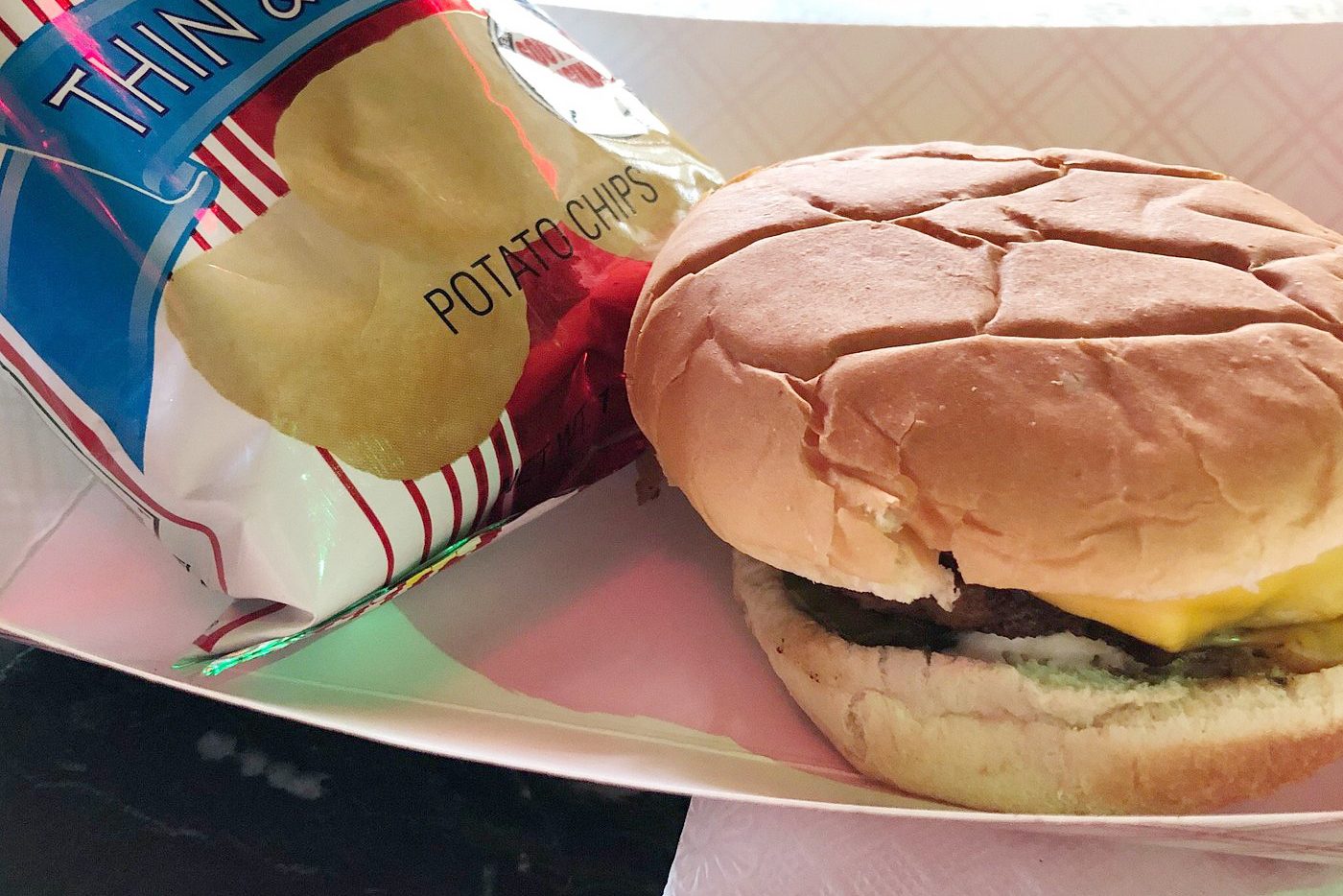 A cheeseburger sits next to a bag of "THIN & CRISPY POTATO CHIPS" in a paper tray on a table.