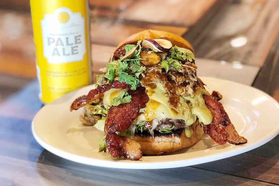 Burger topped with bacon, melted cheese, and herbs sits on a white plate. A yellow can labeled "PALE ALE" stands beside it on a wooden table.