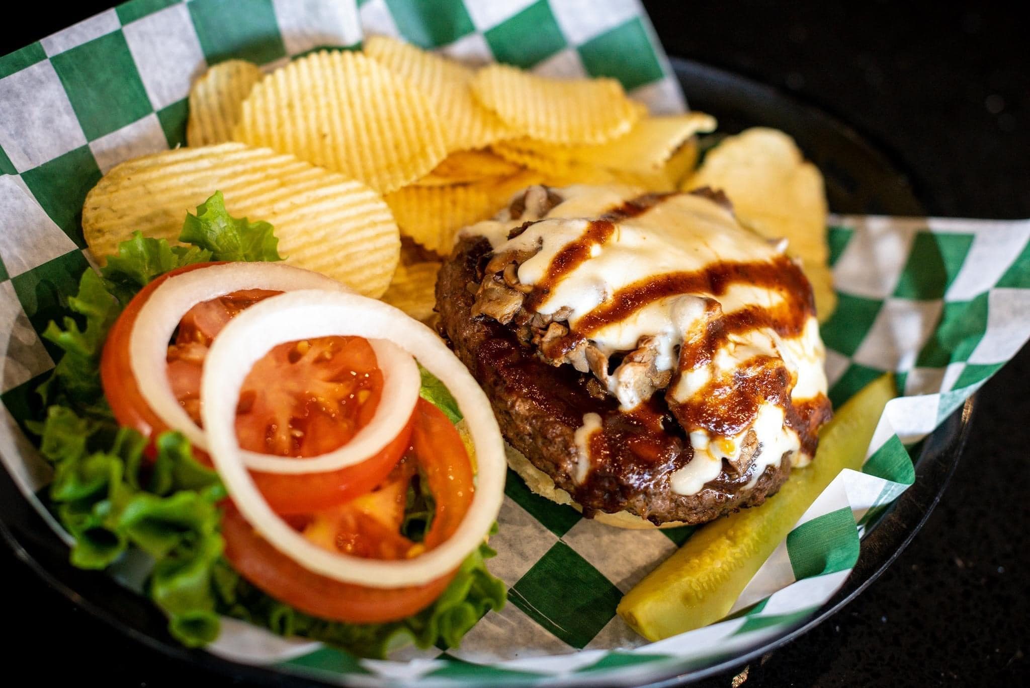 Burger with melted cheese and sauce sits on a bun-less plate, accompanied by crinkle-cut potato chips, lettuce, tomato, onion rings, and a pickle spear.