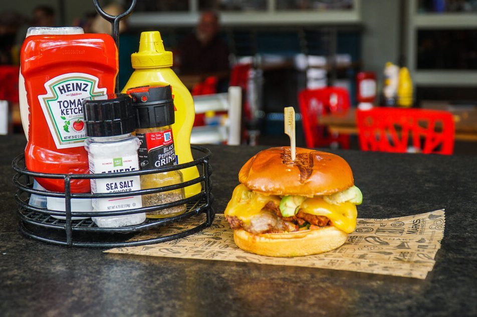 Burger sits on paper next to condiment caddy, including ketchup, mustard, salt, and pepper, on a restaurant table with red chairs in background.