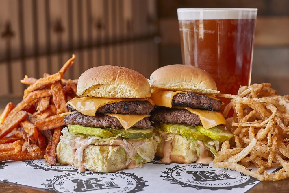 Burgers stacked with cheese and pickles, placed between sweet potato fries and onion rings, sit on branded paper alongside a frothy glass of beer.