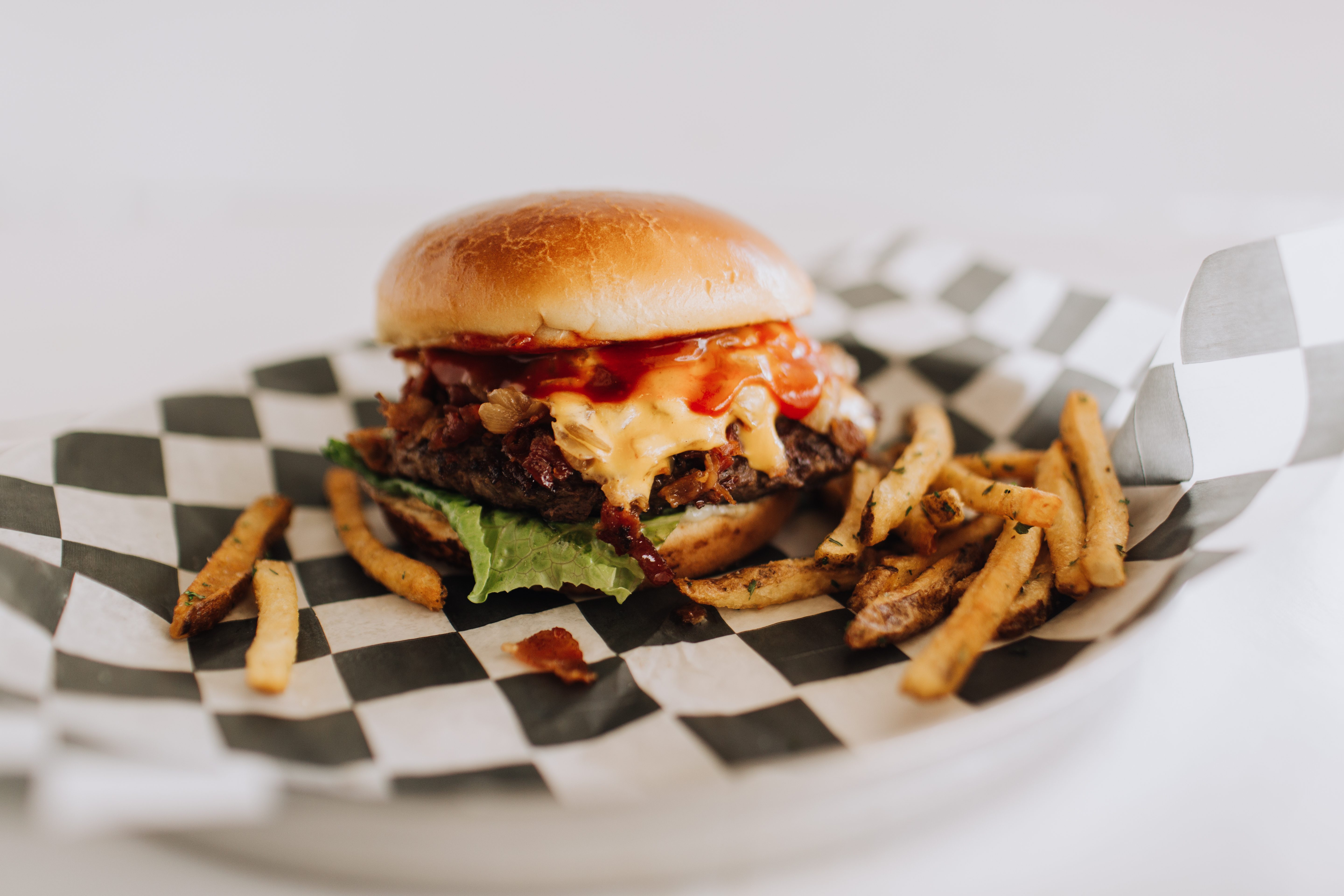 Burger with melted cheese and ketchup sits on checker-pattern paper, surrounded by crispy fries on a white plate.