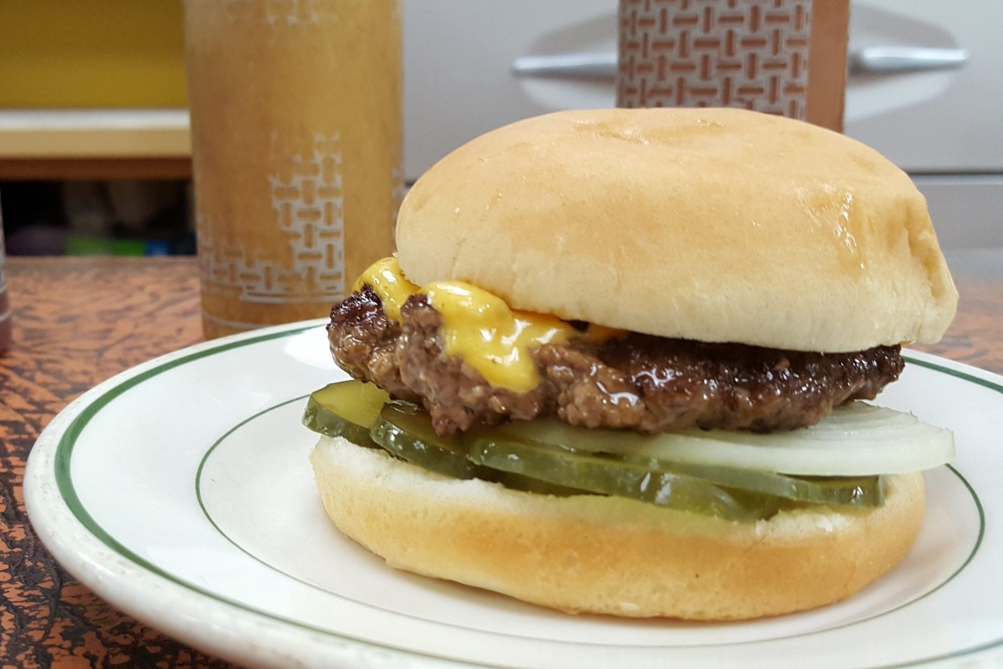 Burger with cheese, onions, and pickles sits on a plate; background shows diner-style setting with condiment bottles.