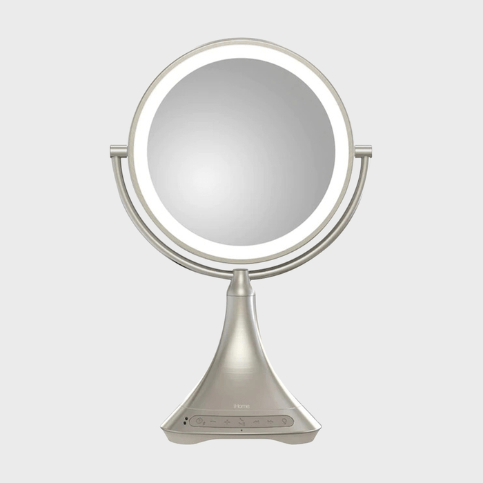 A round mirror emits light, mounted on a stand with controls for power, volume, and Bluetooth, set against a plain background.