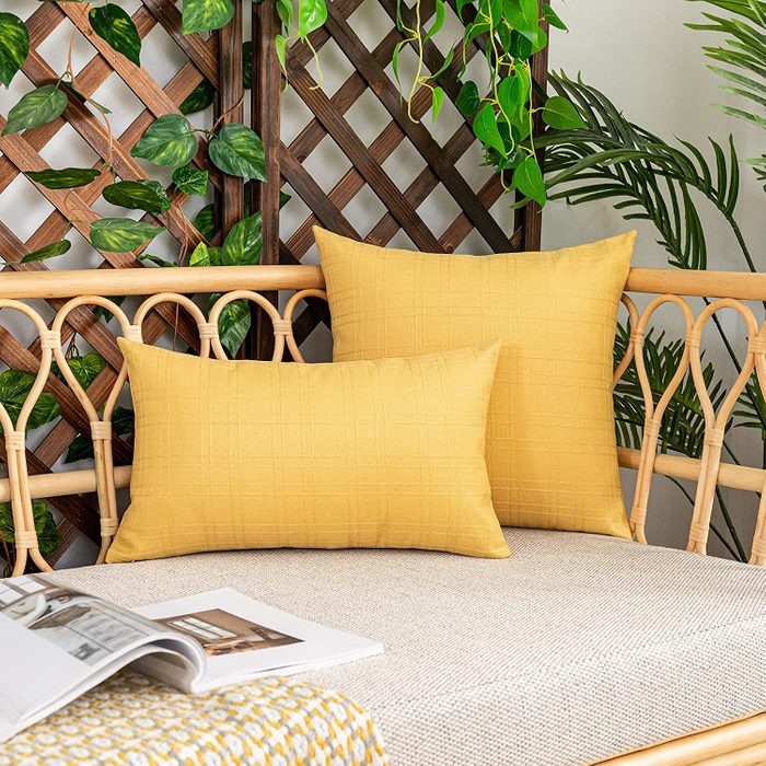 Yellow pillows rest on a wicker bench amid lush green plants, with an open magazine on a patterned blanket nearby.